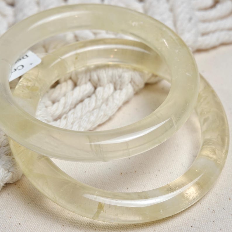 May include: Two clear, translucent bangle bracelets made of polished, light yellow stone. The bracelets are stacked on top of each other.