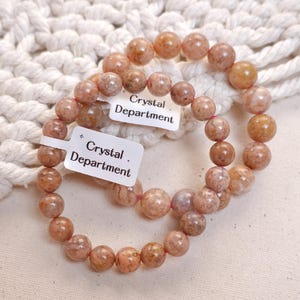 Puede incluir: Dos pulseras de piedras preciosas rosadas y marrones con el texto "Crystal Department" en etiquetas blancas.