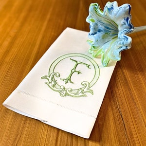 May include: A white linen hand towel with a green embroidered letter "F" inside a decorative circle. A glass flower with blue and green hues rests on the towel. The towel is on a wooden surface.