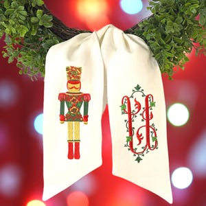 May include: A white fabric sash featuring two embroidered designs. The left side displays a nutcracker figure in red, green, and gold thread. The right side features a red monogram letter U surrounded by green ivy vines and leaves.