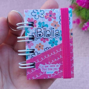 Może przedstawiać: Mały różowo-biały notes w spirali z różową gumką. Okładka zdobiona jest wzorem kwiatowym z brokatem i napisem "Biblia" czarnymi literami. Notes ma srebrny brelok i napis "Lo que viene de Dios trae paz. Juan 14:27" nadrukowany na dole.