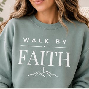 May include: A sage green crewneck sweatshirt featuring white text that reads WALK BY FAITH with a horizontal line and a small dot between the words. Below the text is a simple white line drawing of a mountain range with a cross at the peak.