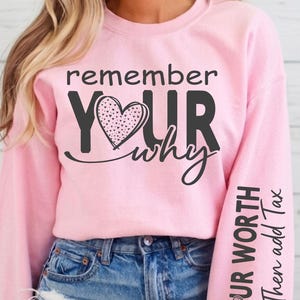 May include: A pale pink sweatshirt featuring the words "remember YOUR why" in black script and a dotted heart design. The right sleeve has the text "KNOW YOUR WORTH Then add Tax". The sweatshirt is worn with blue jeans.