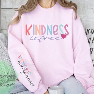 May include: A light pink sweatshirt with the words "KINDNESS is free" in colorful letters and a heart graphic. The sleeve has the words "SPRINKLE it everywhere" in script. The sweatshirt is part of the Pineridge Creators collection.