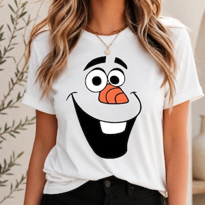 May include: A white short-sleeved t-shirt featuring a large graphic print of a cartoon snowman face. The design includes two round eyes, black eyebrows, a prominent orange carrot nose, and a wide black mouth with a single white tooth.