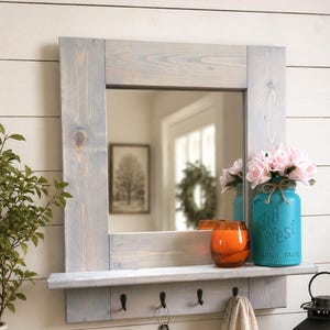 Peut inclure: Un miroir mural rectangulaire avec un cadre en bois teinté gris et une étagère intégrée. L'étagère comporte quatre crochets en métal noir retenant un trousseau de clés et un tissu beige. Un bocal turquoise avec des fleurs roses est posé dessus.