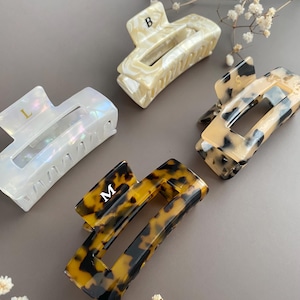 May include: Four hair claw clips in different designs. One is pearlescent white, another is cream, and two are tortoiseshell. Each clip has a letter: L, B, S, and M. The clips are on a grey surface with white flowers.