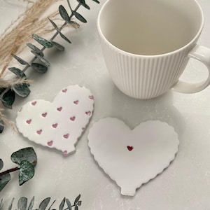 Peut inclure: Deux sous-verres en céramique blanche en forme de cœur. Un sous-verre a un motif de petits cœurs roses. L'autre sous-verre a un seul petit cœur rouge au centre.