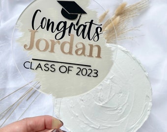May include: A round, clear acrylic cake topper with a gold and white brushstroke design. The topper features the text "Congrats Jordan Class of 2023" and a black graduation cap.