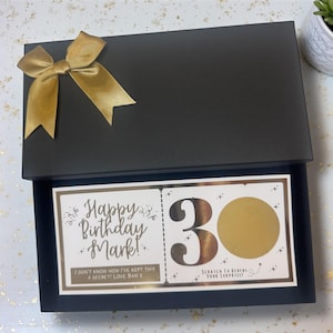 May include: A black gift box tied with a gold ribbon. Inside, a card with the text "Happy Birthday Mark!" and "30" with a scratch-off area. The message "I don't know how I've kept this a secret! Love Sam X" is also printed.