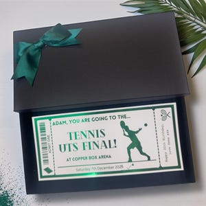 Peut inclure: Un coffret noir orné d'un nœud vert en satin. À l'intérieur, un billet avec le texte "TENNIS UTS FINAL!" et la date "Samedi 7 décembre 2025" est visible. Le billet comprend également le texte "ADAM, YOU ARE GOING TO THE..."