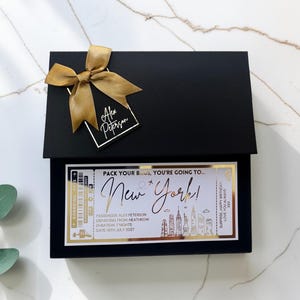 May include: A black rectangular gift box adorned with a gold ribbon and a tag labeled Alex Peterson. The box features a large, gold-foiled boarding pass graphic that reads Pack Your Bags, You're Going To New York, including flight details.