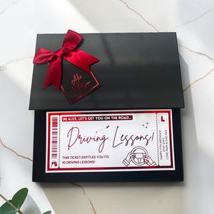 May include: A black gift box featuring a red satin bow and a matching tag. Inside, a white ticket with red accents reads: Alex, let's get you on the road... Driving Lessons! This ticket entitles you to: 10 driving lessons! Happy 17th birthday.