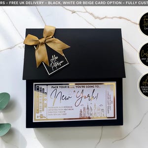 May include: A black rectangular gift box featuring a gold ribbon and a tag labeled Alex Peterson. A boarding pass style card is attached, reading Pack your bags, you're going to New York, with passenger details, a skyline graphic, and gold foil accents.