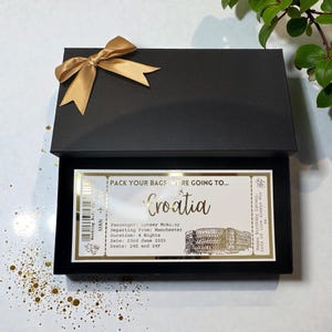 May include: A black gift box tied with a gold ribbon, displaying a decorative ticket. The ticket, with gold text, says "Pack Your Bags, We're Going To... Croatia". It includes passenger details and the date of travel.
