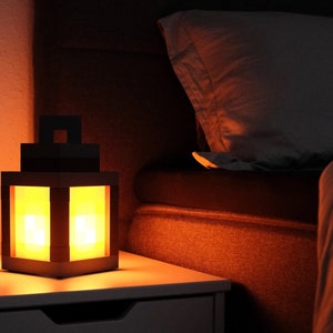 May include: A wooden lantern with a warm, orange glow illuminates a bedside table. The lantern has a square shape with a clear glass panel on each side. The lantern is sitting on a white nightstand with a drawer.
