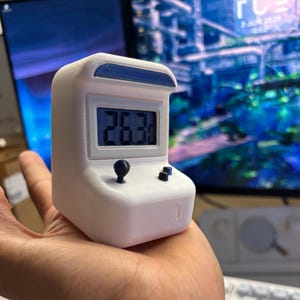 Puede incluir: Un reloj digital blanco en miniatura con forma de máquina recreativa, con una pantalla azul que muestra la hora. Tiene una parte superior azul, un joystick negro y dos botones negros. El reloj se sostiene en una mano.