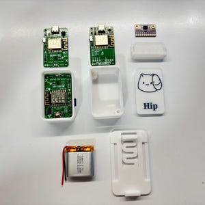 Puede incluir: Un surtido de componentes electrónicos, que incluyen placas de circuito verde, carcasas de plástico blanco, una pequeña batería y un componente con la inscripción "Hip" y un diseño de gato de dibujos animados. Los componentes están dispuestos sobre una superficie blanca.