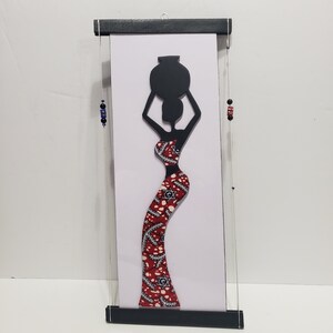 May include: Black silhouette of a woman carrying a pot on her head, wearing a red and white patterned dress. The silhouette is mounted on a white background and framed with a black wooden frame.