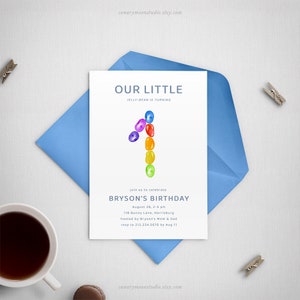 以下が含まれることがあります： 青い文字で書かれた白い招待状。カードには「Our Little Jelly Bean is Turning 1」と虹色のグミのデザインが書かれています。また、「Join us to celebrate Bryson's Birthday」という文言と、日付、時間、場所、RSVPに関する情報が記載されています。