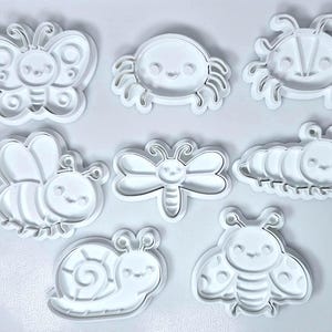 Pode incluir: Uma coleção de cortadores de biscoitos de plástico branco em várias formas de insetos, incluindo uma borboleta, caranguejo, besouro, abelha, libélula, lagarta, caracol e joaninha. Cada cortador tem um rosto sorridente.