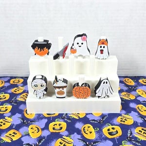 May include: A white tiered display holding various Halloween-themed figurines. The figurines include a cauldron, a knife, ghosts, a mummy, a coffee drink, a pumpkin, and a bat. The display sits on a blue fabric with a pumpkin and bat pattern.