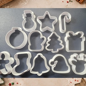 Op de afbeelding: Een set van elf witte plastic koekjesvormen in diverse feestelijke vormen, waaronder een sneeuwpop, ster, zuurstok, kerstbal, want, kerstboom, cadeau, peperkoekmannetje, kerstsok, bel, kerstmuts en een rendierkop.