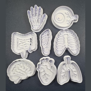 Peut inclure: Un ensemble de huit emporte-pièces en plastique blanc en forme de parties anatomiques humaines, incluant une main, un œil, des intestins, un pied, une cage thoracique, un cerveau, un cœur et des poumons, disposés sur une surface gris foncé.