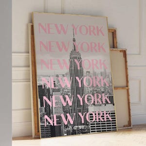 May include: A framed print featuring a black and white photograph of the New York City skyline, with the Empire State Building prominently displayed. The word "NEW YORK" is repeated in pink across the image. The frame has a gold border.