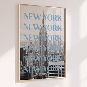May include: Framed print featuring a black and white photograph of the New York City skyline, with the Empire State Building prominently displayed. The word "NEW YORK" is repeated in light blue vertically across the image. The frame is gold.