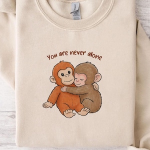 May include: A beige sweatshirt featuring a cartoon illustration of two hugging monkeys. The text "You are never alone" is above the monkeys in brown lettering. The sweatshirt is made of a soft material.