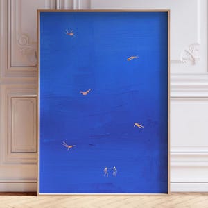 May include: A framed abstract art print featuring a deep blue background with several small, golden-colored figures. The figures appear to be swimming or floating. The artwork is displayed in a room with white walls and a wooden floor.