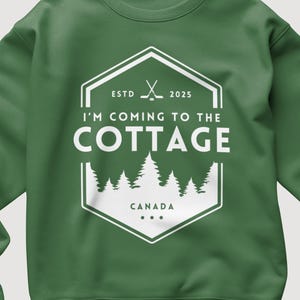 May include: Green sweatshirt with a white graphic that reads "I'M COMING TO THE COTTAGE" inside a hexagon with crossed hockey sticks and the text "CANADA". The sweatshirt has a crew neck and long sleeves.