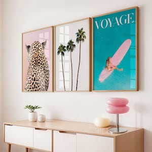 May include: Three framed art prints. The first features a leopard's spotted back against a pink background. The second shows palm trees. The third has a person on a pink surfboard in turquoise water, with the word "VOYAGE".