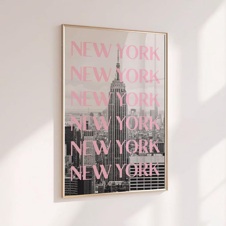 May include: Framed print featuring a black and white photograph of the New York City skyline, with the Empire State Building prominently displayed. The word "NEW YORK" is repeated in pink, vertically across the image. The frame is gold.