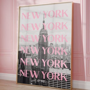 May include: A framed print featuring a black and white photograph of the New York City skyline, with the Empire State Building prominently displayed. The word "NEW YORK" is repeated in pink over the image. The frame is gold.