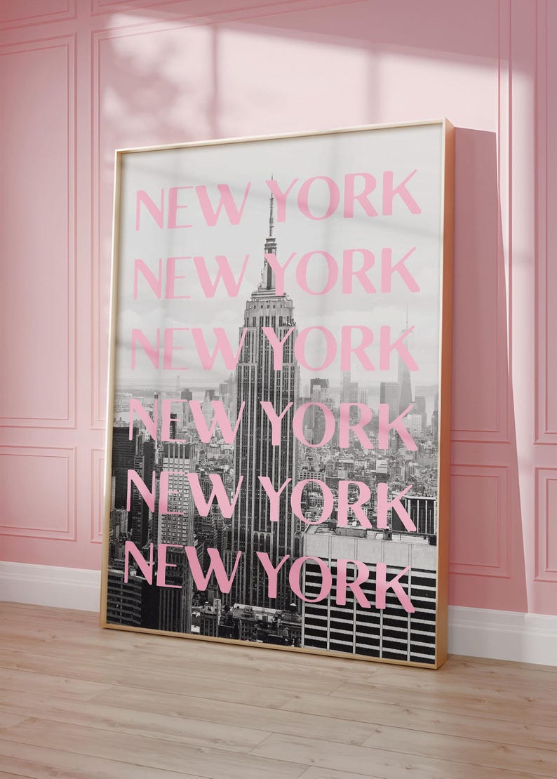 May include: A framed print featuring a black and white photograph of the New York City skyline, with the Empire State Building prominently displayed. The word "NEW YORK" is repeated in pink over the image. The frame is gold.