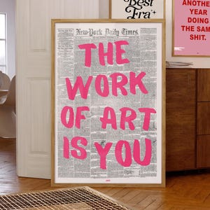 Puede incluir: Una impresión enmarcada con un fondo de periódico y texto rosa que dice "The Work of Art is You".