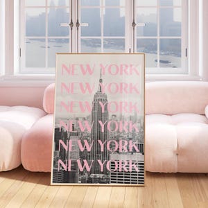 May include: A framed print featuring a black and white image of the New York City skyline, with the Empire State Building prominently displayed. The word "NEW YORK" is overlaid in pink, repeated vertically. The print is displayed in a room with a pink sofa.