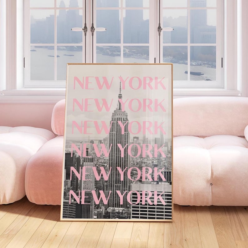 May include: A framed print featuring a black and white image of the New York City skyline, with the Empire State Building prominently displayed. The word "NEW YORK" is overlaid in pink, repeated vertically. The print is displayed in a room with a pink sofa.
