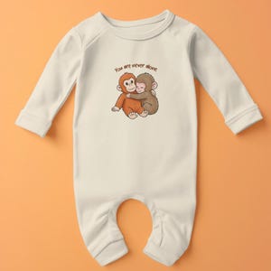May include: Cream-colored baby onesie with long sleeves and a round neckline. The onesie features an illustration of two hugging monkeys with the text "You are never alone" above them. The monkeys are brown and orange.