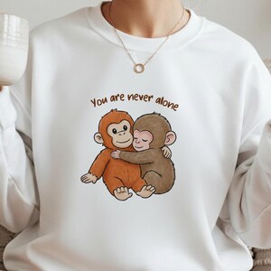May include: White sweatshirt featuring a cartoon illustration of two hugging monkeys in shades of brown and orange. The text "You are never alone" is above the monkeys in brown lettering.