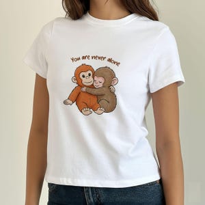 May include: White t-shirt featuring a cartoon illustration of two hugging monkeys. The monkeys are brown and orange, with the text "You are never alone" above them. The shirt is a crew neck style.