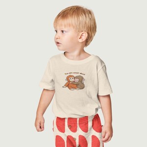 May include: A cream-colored t-shirt featuring a graphic of two hugging monkeys. The text "You are never alone" is above the monkeys. The child is wearing red patterned shorts.