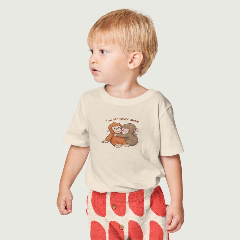 May include: A cream-colored t-shirt featuring a graphic of two hugging monkeys. The text "You are never alone" is above the monkeys. The child is wearing red patterned shorts.