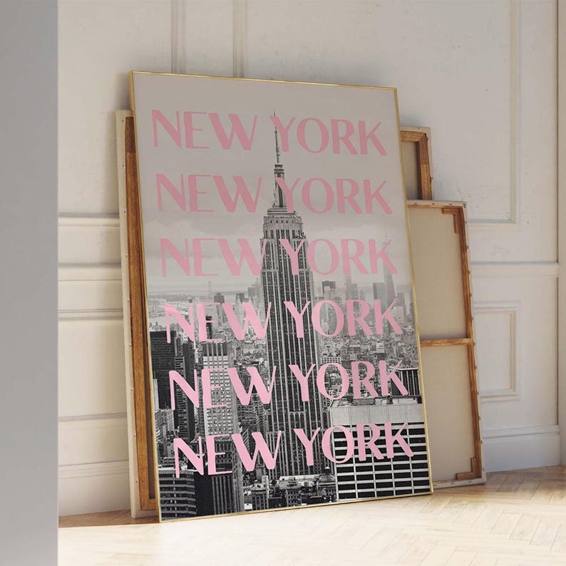 May include: A framed print featuring a black and white photograph of the New York City skyline, with the Empire State Building prominently displayed. The word "NEW YORK" is repeated in pink across the image. The frame has a gold border.