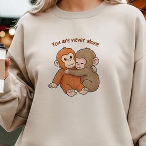 May include: Beige sweatshirt featuring a graphic of two hugging cartoon monkeys. The monkeys are brown and orange, with the text "You are never alone" above them in brown lettering. The sweatshirt is a neutral color.