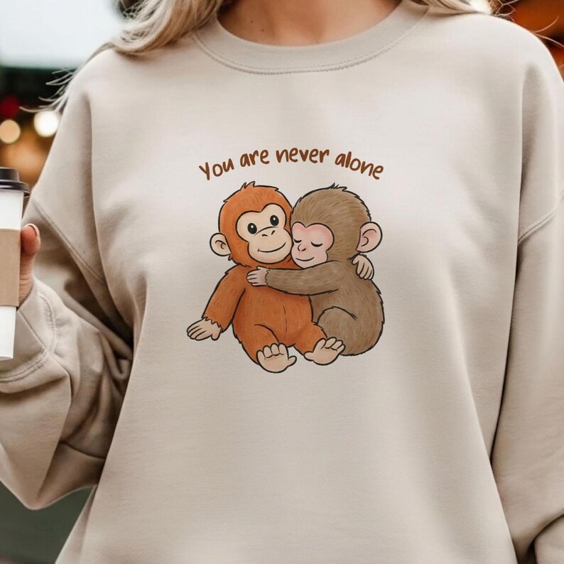 May include: Beige sweatshirt featuring a graphic of two hugging cartoon monkeys. The monkeys are brown and orange, with the text "You are never alone" above them in brown lettering. The sweatshirt is a neutral color.