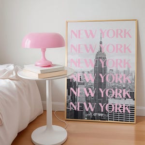 May include: A pink mushroom-shaped lamp sits on a white side table with books. A framed black and white photo of the New York City skyline with the text "NEW YORK" in pink is behind the table. The room has a minimalist design.