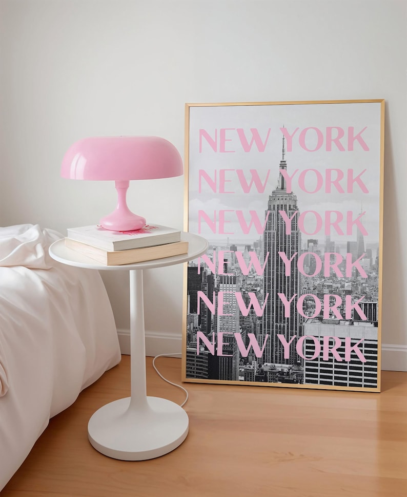 May include: A pink mushroom-shaped lamp sits on a white side table with books. A framed black and white photo of the New York City skyline with the text "NEW YORK" in pink is behind the table. The room has a minimalist design.